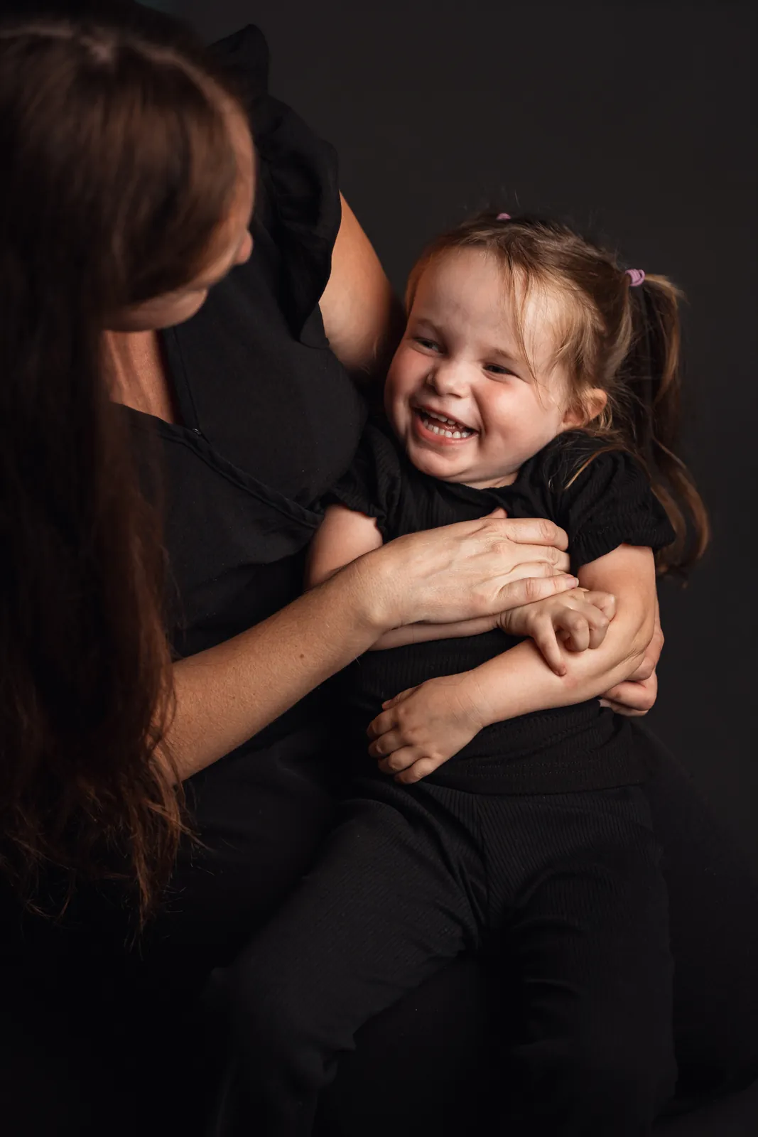 Athena éclate de rire, assise sur les genoux de sa maman Megan qui la tient dans ses bras, sur fond sombre