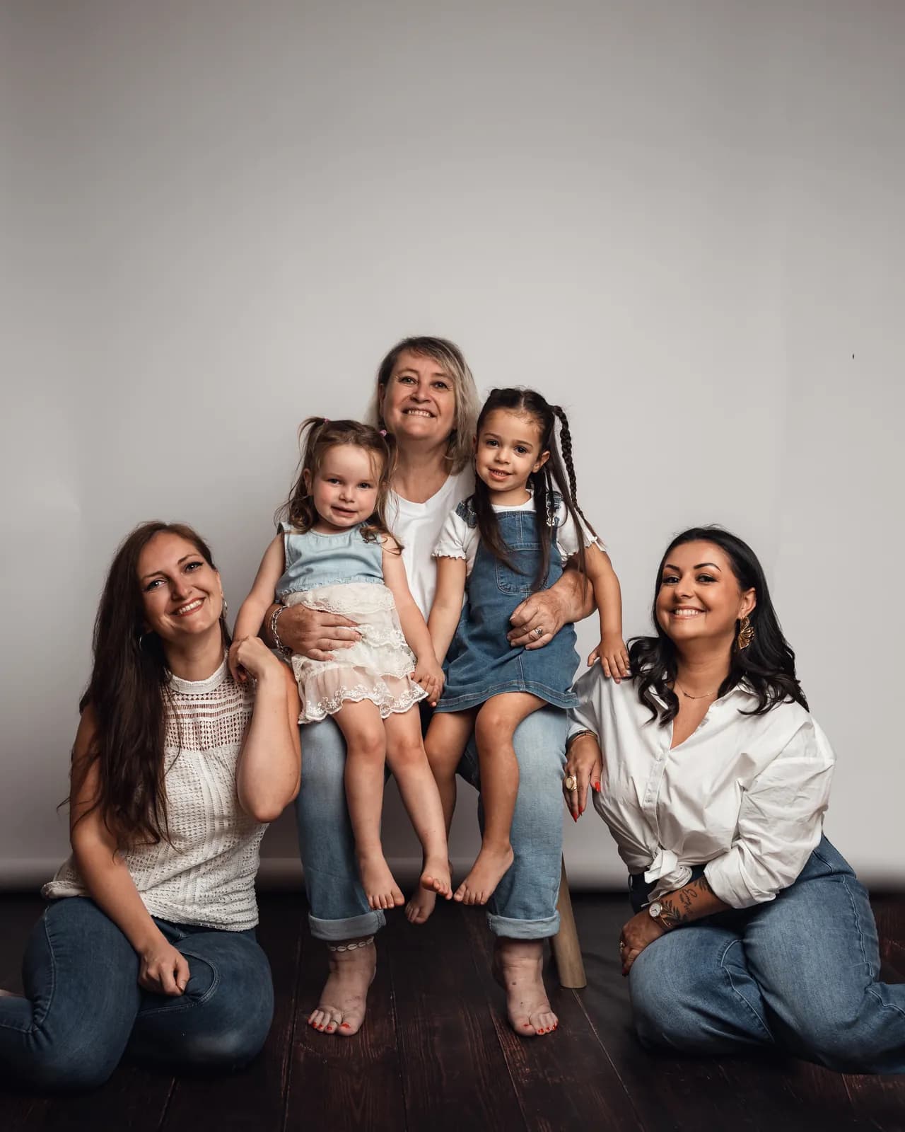 La grand-mère assise sur un tabouret avec ses deux petites-filles contre elle, ses deux filles agenouillées de chaque côté, en blanc et en jean sur fond clair