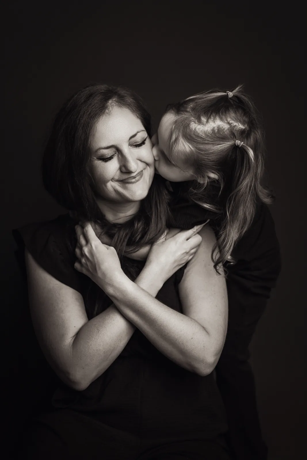 En noir et blanc, Athena fait un bisou sur la joue de sa maman Megan, qui sourit les yeux fermés, les bras croisés sur ceux de sa fille