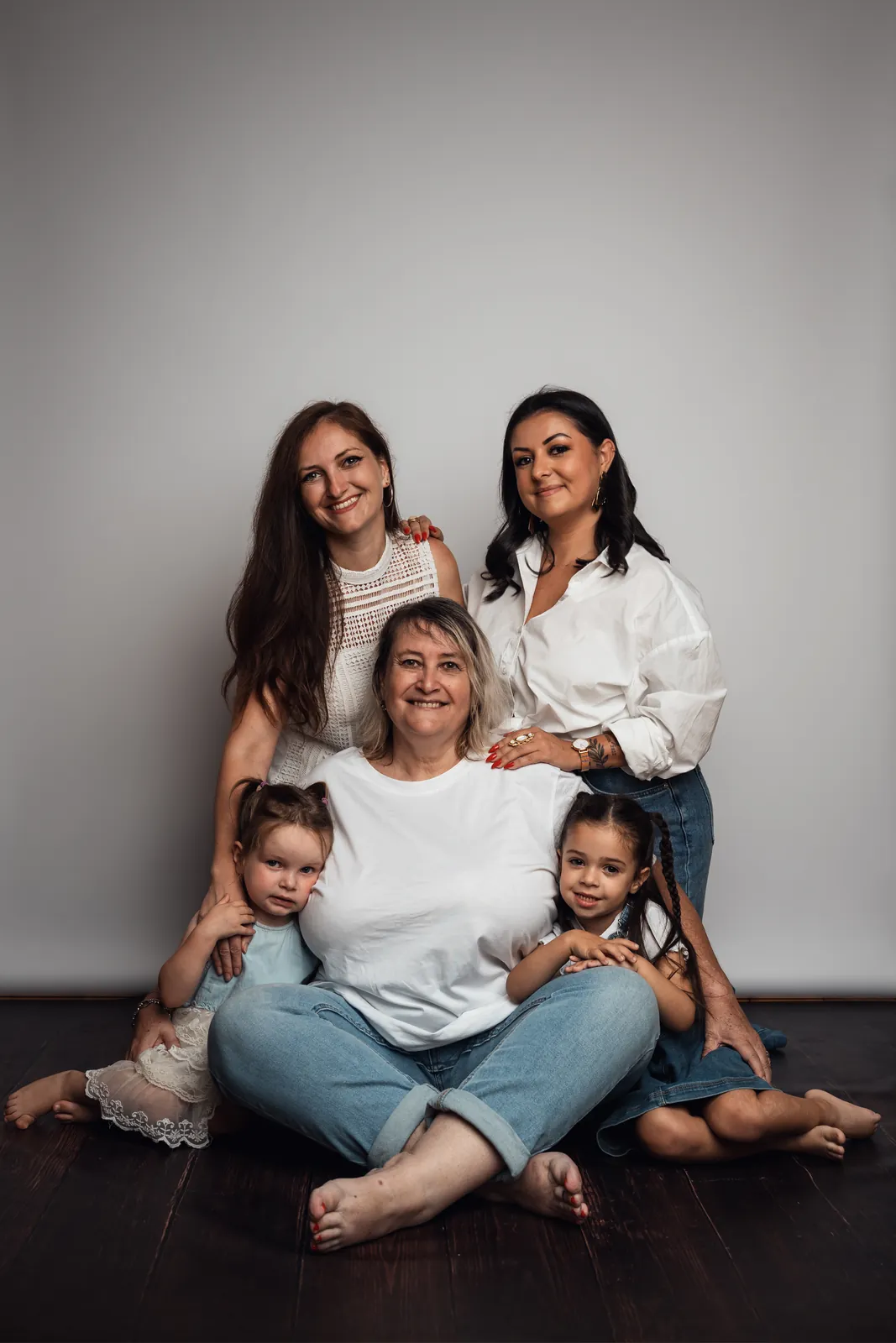 Une grand-mère assise en tailleur sur le parquet du studio, entourée de ses deux filles debout et de ses deux petites-filles, toutes en blanc et en jean