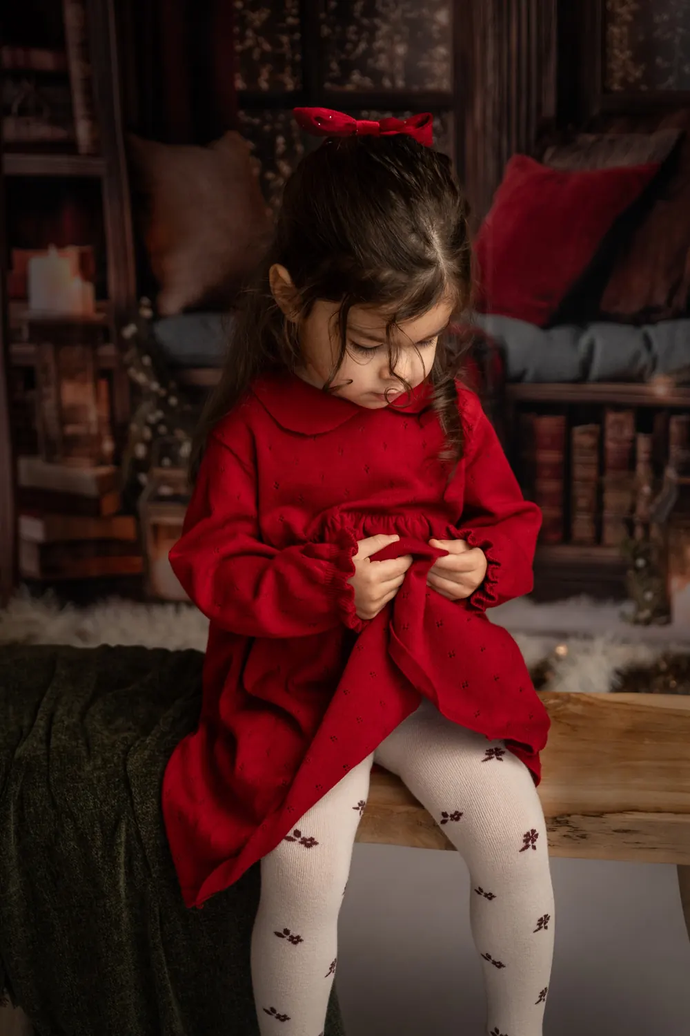 Petite fille en robe rouge assise sur un banc, regard vers le bas, décor de Noël rouge et vert